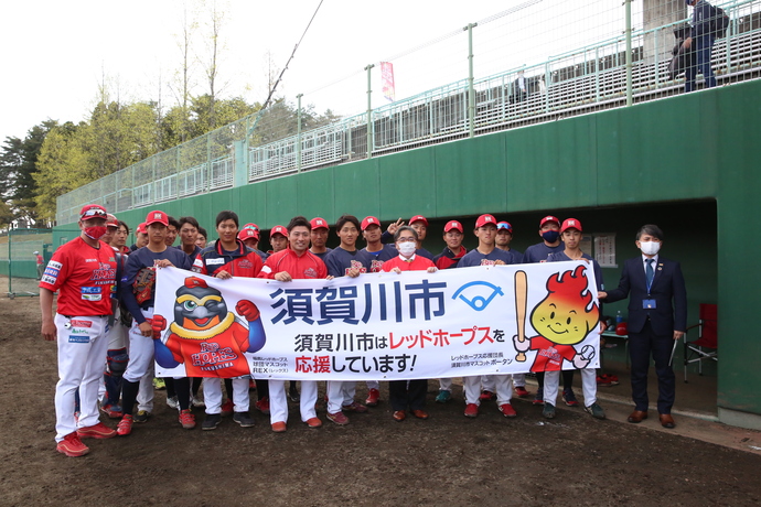 赤いユニフォームと白いズボンを着用した野球チームの選手たちと、安藤副市長、スーツ姿の男性を含む関係者一同が、「須賀川市はレッドホープスを応援しています！」と書かれた横断幕を持ってグラウンドに立っている集合写真