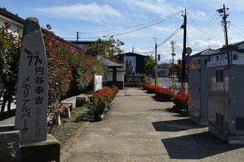 「77 円谷幸吉メモリアルパーク」と刻まれた石碑が左手前に立ち、赤い花の鉢植えが並ぶ歩道の先に、モニュメントと大きな写真パネルの展示が見える、円谷幸吉メモリアルパークの写真