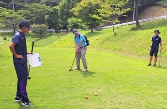緑の芝生が広がるパークゴルフ場で、男性二人が見守る中、1人の男性がゴルフクラブを使ってボールを打とうとしている瞬間の写真