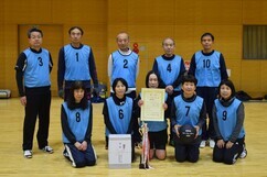 体育館にて、水色のゼッケンを着用した男女10名のチームメンバーが、手にした賞状やバレーボール、トロフィー、そして白い副賞の箱を囲み、達成感のある表情で二列に並んで撮影された大会の記念写真