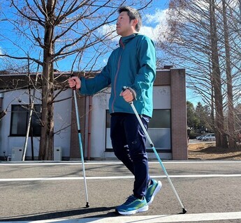 冬枯れの木々に囲まれた建物沿いの道路で、水色の上着を着た男性が、両手に白と水色の配色のポールを持って歩いている、ポールウォーキングの様子の写真