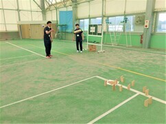 屋内のスポーツ施設にて、競技エリアの奥に立てられた複数の木製ピンを目がけて、手前の男性がバトン状の木をアンダーハンドで正確に投げようとしている、モルック競技に真剣に取り組む様子を捉えた風景写真