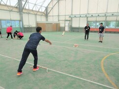 屋内のスポーツ施設にて、手前の男性が木製のバトンを前方の標的に向かって投じ、それを見守る周囲の参加者たちが競技の行方を注視している、フィンランド発祥のスポーツであるモルックを楽しんでいる風景写真