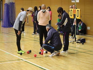 体育館のコート上に集まった赤、青、白の複数のボールを囲むように、審判員やジャージ姿の参加者たちが集まり、得点盤の横でボールの配置を真剣な表情で確認している、ボッチャの試合中の一コマを捉えた写真