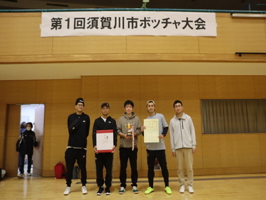 「第1回須賀川市ボッチャ大会」と書かれた横断幕が掲げられた体育館の壁を背景に、優勝したチームの男性5名がトロフィーや賞状、副賞の箱を誇らしげに手に持って並び、達成感のある笑顔を見せている記念写真