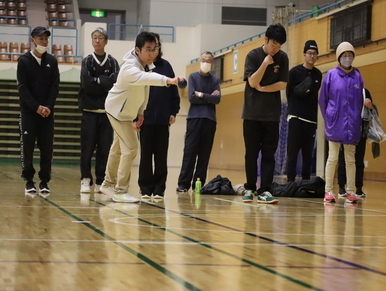 体育館のような屋内で、白いパーカーを着用した男性が真剣な表情で黒い球を投げ、その様子を後方でマスクをした男女数人の参加者たちが見守っている、ボッチャ競技を楽しんでいる写真