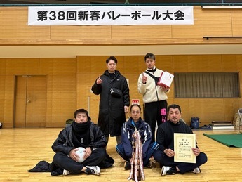 「第38回新春バレーボール大会」の横断幕が掲げられた体育館で、賞状やトロフィー、記念品を手にした5名の男性たちが、大会の成果を祝うように一列に並んでカメラに微笑みかけている写真