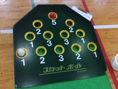 体育館の床に置かれた「スカットボール」という競技用の緑色のマットに、得点となる1から5の数字が振られた複数の穴が開いており、そのうち5点の穴に赤いボールが、1点の穴に白いボールがそれぞれ入っている様子を真上から捉えた写真