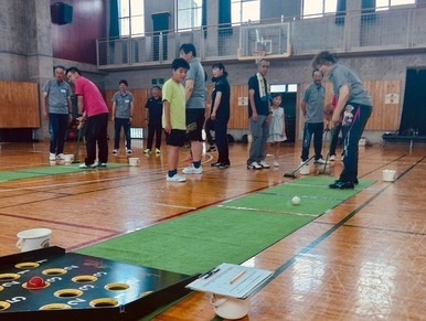 体育館のコートに敷かれた長い緑色のマットの上で、高齢の女性や子どもを含む幅広い世代の市民が、スティックを使いボールを穴に入れて得点を競うスカットボールという軽スポーツを和気あいあいと楽しんでいる写真