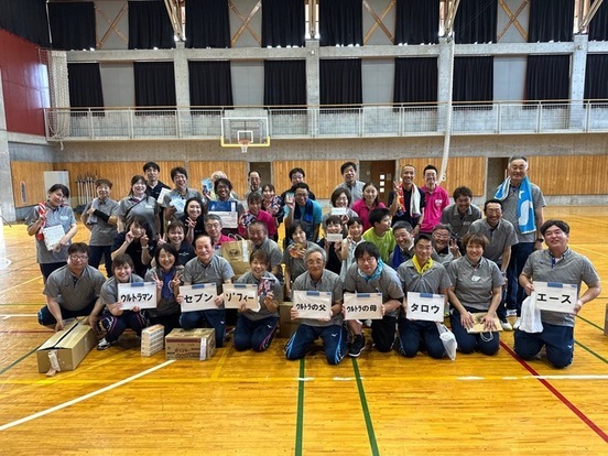 体育館のコート上で、お揃いのグレーのポロシャツや色とりどりのウェアを着た老若男女約30名の参加者たちが、「ウルトラマン」「セブン」「タロウ」など歴代ヒーローの名が書かれたパネルを掲げて笑顔で集合した記念写真
