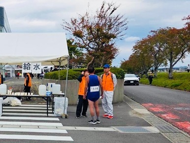 マラソン大会のコース沿いに設置された給水所で、白地に黒文字で「給水」と書かれた案内が掲げられたテントの横に、青いユニフォームのランナーとオレンジ色のベストを着た運営スタッフが立ち、話をしている写真