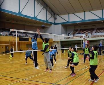 広い体育館の中で、ネットを挟んで水色のユニフォームと黄緑色のビブスを着用した選手たちが、高く跳び上がり空中のボールを競り合う白熱したラバーバレーボールの試合風景を捉えた写真