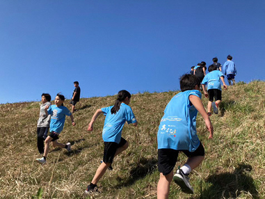 屋外で草の生えた斜面を走って上り下りするトレーニングを行っている子どもたちの写真