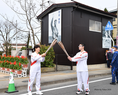 円谷幸吉選手の肖像が掲げられた建物の前で、白いユニフォームを着た男女のランナーが聖火リレーのトーチを掲げて交差させる「トーチキス」を行っている、東京2020オリンピックの聖火リレーの瞬間を捉えた写真