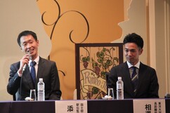 テーブルに並んで座った、マイクを手に笑顔で話す男性と、その隣で静かに耳を傾けるスーツ姿の男性の写真