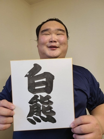 力士の白熊関が、自身の四股名である「白熊」という文字が力強く墨で書かれた正方形の色紙を両手で掲げ、目を細めて穏やかに微笑んでいる写真