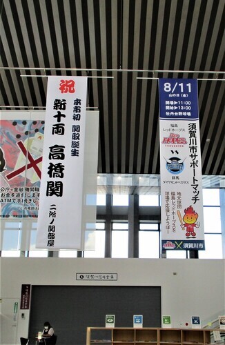 高い天井から、「祝新十両高橋関」と記された昇進を祝う大きな垂れ幕と、その隣に「須賀川市サポートマッチ」の開催を告知するポスター風の垂れ幕が並んで吊るされている、市庁舎「みんなのスクエア」のロビーの広々とした吹き抜け空間の写真