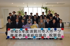 「令和5年度須賀川市スポーツ推進委員連絡協議会祝賀会」と記された横断幕を前に、フォーマルな服装の男女が三列に整列し、中央の出席者2名が花束を抱えて記念撮影を行っている集合写真