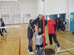 体育館にて、赤い支柱にバレーボールネットを張り、高さの調整や固定作業を協力して行っている、ジャージ姿の男女や赤いポロシャツを着用したスタッフたちの準備風景を記録した写真