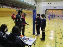 体育館にて、青い支柱カバーが巻かれたバレーボールネットの横で、ジャージ姿の審判員やスタッフたちが試合開始前の確認や打ち合わせを真剣な表情で行っている、大会運営の様子を捉えた写真
