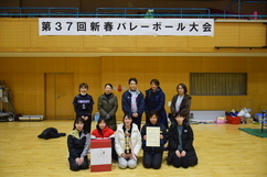 「第37回新春バレーボール大会」の横断幕が掲げられた体育館で、賞状やトロフィー、副賞の箱を囲みながらユニフォームや防寒着姿の女性チームメンバーが笑顔で整列して撮影された、大会の成果を祝う記念写真