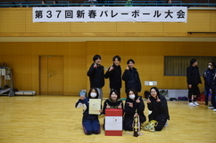 「第37回新春バレーボール大会」の横断幕が掲げられた体育館で、前列に座る女性たちが賞状やトロフィー、大きな副賞の箱を囲み、後列の男女混合メンバーと共に笑顔やポーズで勝利を喜んでいる様子を写した記念写真