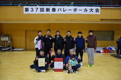「第37回新春バレーボール大会」と書かれた横断幕の下で、賞状やトロフィーを手に持ち笑顔で集合写真に応じる、ユニフォームやジャージ姿の男性選手たちと子供が体育館のコートに並んでいる記念写真
