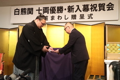「白熊関十両優勝・新入幕祝賀会化粧まわし贈呈式」と書かれた看板の下、金屏風の前で紋付き袴姿の白熊関が、スーツ姿の男性から目録のような品を恭しく受け取っている、お祝いの席での記念すべき瞬間を捉えた写真