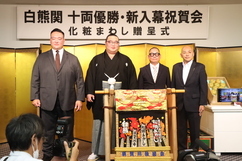 「白熊関十両優勝・新入幕祝賀会」の会場で、紋付き袴姿の白熊関を囲んでスーツ姿の男性たちが金屏風の前に並び、手前には贈呈された華やかな化粧まわしが誇らしげに飾られている、記念すべき贈呈式の様子を捉えた写真