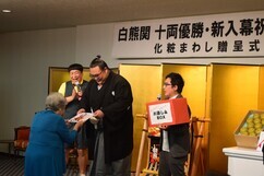 「白熊関十両優勝・新入幕祝賀会」の贈呈式にて、金屏風の前で紋付き袴姿の白熊関が笑顔で高齢の女性に記念品を手渡しており、横では赤い箱を持った司会者やゲストが温かく見守っている、和やかな交流の場面を捉えた写真