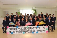 「令和6年度須賀川市スポーツ推進委員連絡協議会祝賀会」と書かれた看板の前で、スーツや礼服に身を包んだ大勢の出席者が並び、前列の人々が色鮮やかな花束を抱えてにこやかに撮影に応じている記念写真