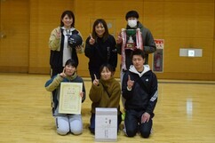 体育館にて、賞状や優勝カップ、バレーボール、そして白い副賞の箱を囲んだ男女6名のチームメンバーが、人差し指を立てるナンバーワンのポーズや笑顔でカメラに向かって並んでいる、大会の勝利を祝う記念写真