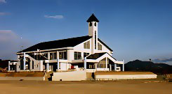 山々を遠景に望む広々とした敷地に白を基調とした壁面に黒い大屋根が載り、中央には時計台のような細長い塔がそびえ立つ特徴的なスポーツ施設の全景を捉えた写真