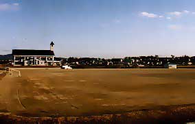 遮るもののない青空と遠くの街並みを背景に、左奥には小さな塔のような造形を持つ2階建ての建物が佇む広々とした土のグラウンドが広がったいる写真