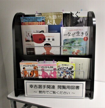「幸吉選手関連 閲覧用図書 ～館内でご覧ください～」の案内が貼られたマガジンラックに様々な本屋雑誌が並んでいる写真