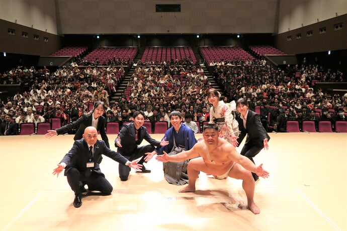 広いホールのステージの上で、市長とまわし姿のあかつさん、5人の実行委員の男女が両腕を広げたポーズをとり、観客席に座った大勢の出席者と一緒に写った記念写真