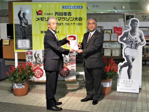 「円谷幸吉メモリアルマラソン大会」の大きな看板や等身大パネルが設置された前で、スーツ姿の安藤会長と橋本市長並んで立ち、目録の封筒を両側から持っている記念写真