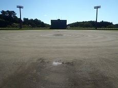 奥に2本の照明とその中央の得点ボードが見える土のグラウンドの写真
