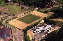 緑豊かな山々に囲まれた平坦な土地に、中央の長方形のグラウンド、左上の野球場、そして手前に特徴的な屋根を持つモダンな平屋建ての施設と広い駐車場が整備されている様子を、空から斜めに俯瞰して捉えた、施設の全体像を把握できる航空写真