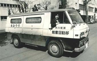 建物の前に、移動図書館うつみねと書かれたワゴン車が停車し、運転席の人物が窓から顔を出し笑顔でカメラの方を見ている白黒写真