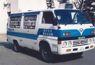 停車している白い車体に青の線が入った移動図書館のワゴン車の窓から、たくさんの本が並んだ本棚が見えている写真