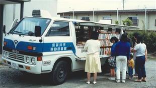 停車している白い車体に青の線が入った移動図書館のワゴン車の側面棚が大きく開いて、本がぎっしり並んだ本棚の前に、数名の大人と子どもが集まり本を選んでいる様子の写真