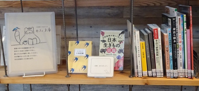 木製の棚に本や案内板が並んでおり、左側には「カフェ文庫」と書かれたイラスト入りの案内板が立てられ、その隣には複数の書籍が並んでいる店舗内での設置例の写真