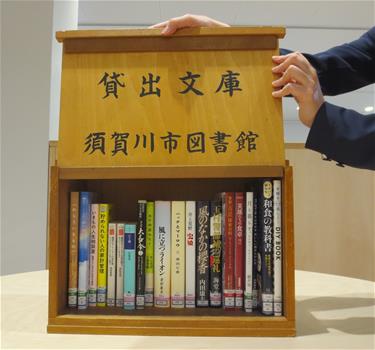 木製の箱型書棚の、「貸出文庫 須賀川市図書館」と黒字で刻印された側面を上にスライドして開き、棚の中に約20冊の様々な背表紙の本がぎっしりと並べられている様子を捉えた写真