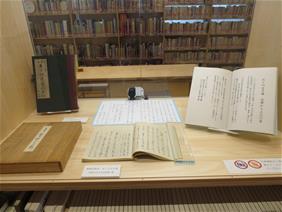 展示ケース内に古書や資料が丁寧に配置され背景の書架とともに落ち着いた雰囲気で示されている写真