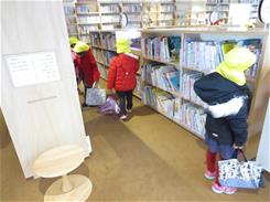 明るい図書館で黄色い帽子の子どもたちが本棚を行き来しバッグを手に本を選んでいる様子の写真