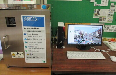金属製の除菌BOXとパソコンモニターが机上に並び、案内表示と機器類が整然と配置された写真