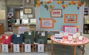 ブドウの葉の飾り付けが施された「読書週間」の展示コーナーで、年齢別におすすめの本が入ったカラフルな4種類のバッグが並び、丸いテーブルの上に複数の本が展示されている様子の写真