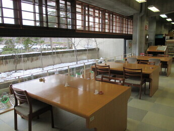 大きな窓から外の景色を望む図書館の静かな閲覧席に木製の机と椅子が整然と並ぶ写真
