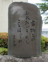 「書物との ふれあいうれし ことはじめ まさお」と刻まれた、光沢のある濃い灰色の句碑の写真